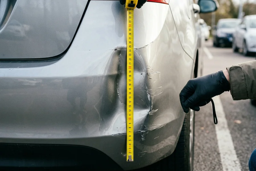 A close-up view of a metallic car bumper dent being measured with a yellow tape measure.